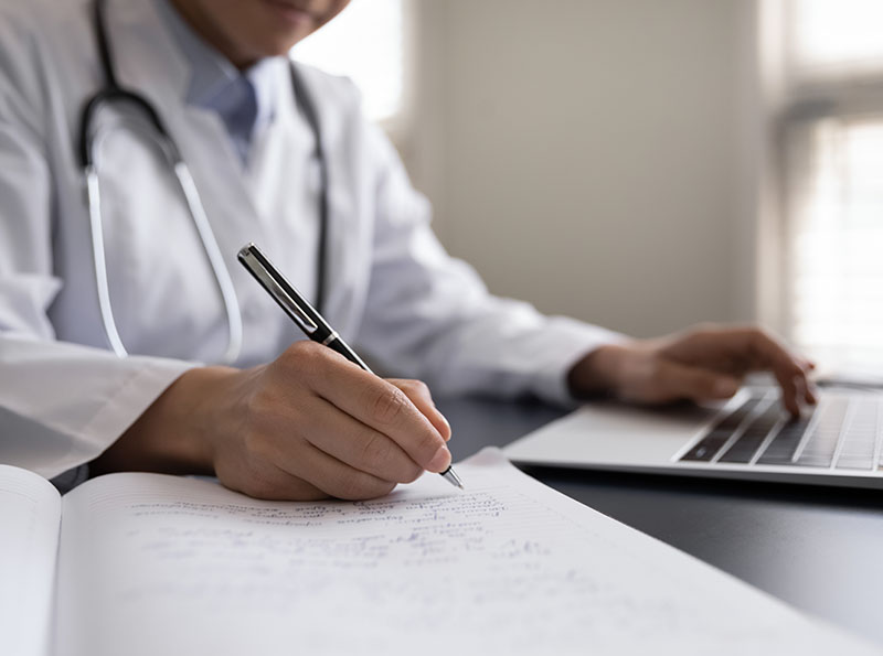 Crop close up Indian woman doctor in white uniform with stethoscope taking notes, using laptop, writing in medical journal, professional therapist practitioner filling documents or patient card